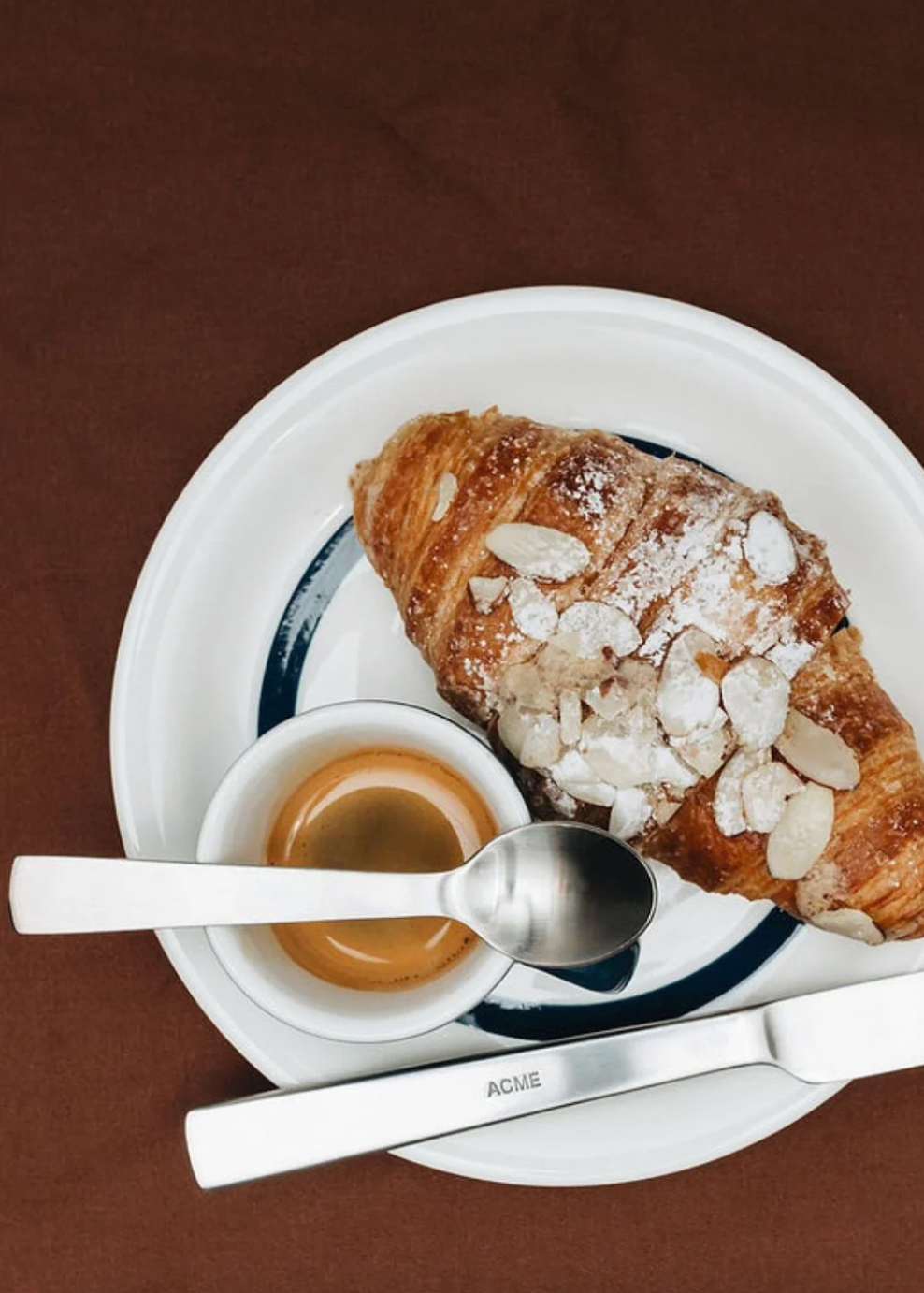 Croissant with almonds and a cup of coffee on a plate with a spoon, all branded 'ACME'.
