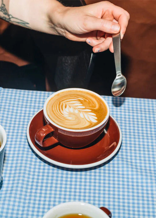 Coffee with latte art on a checkered tablecloth, with a hand holding a spoon.