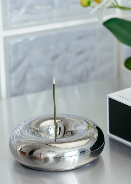 Silver incense burner with a lit incense stick on a reflective surface.