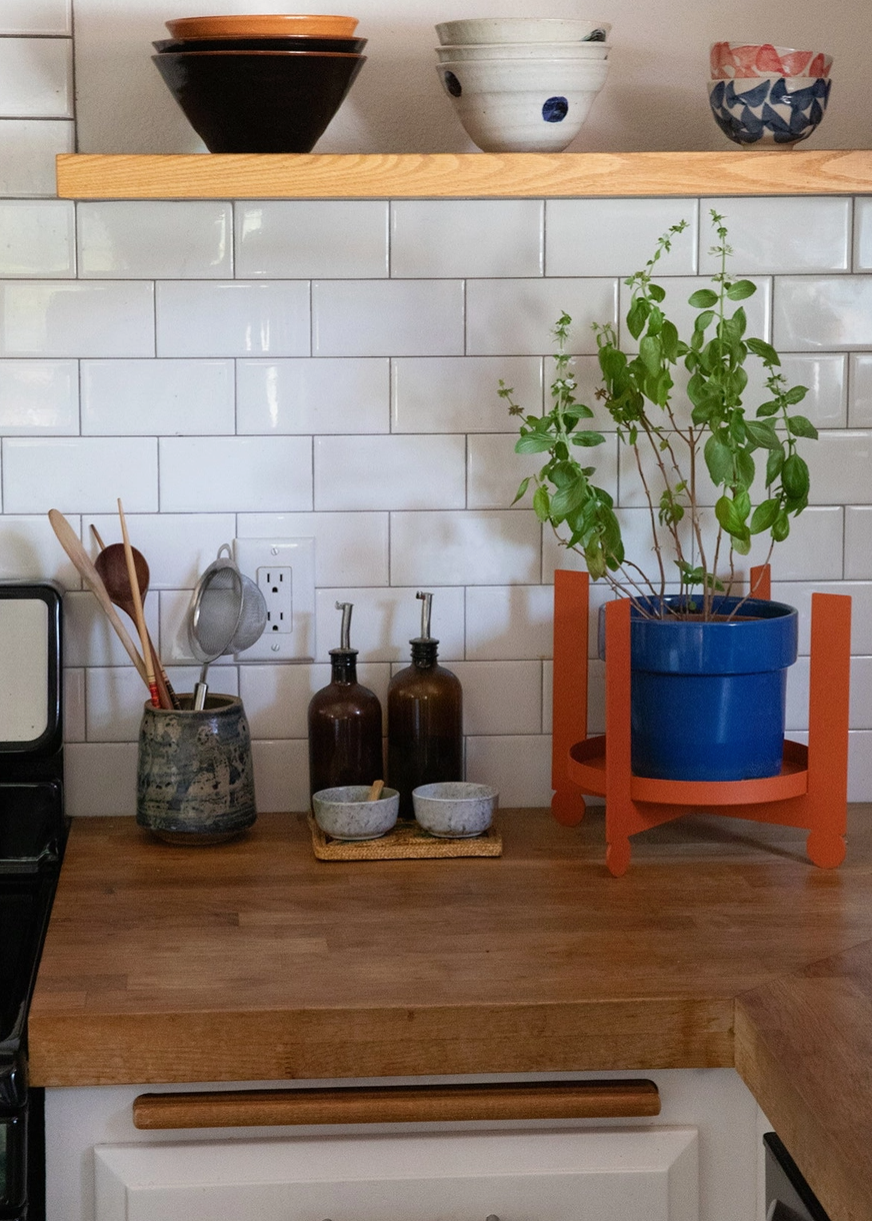 A wooden kitchen counter with a potted plant on an orange metal plant stand, oil bottles, and a pitcher with kitchen utensils.