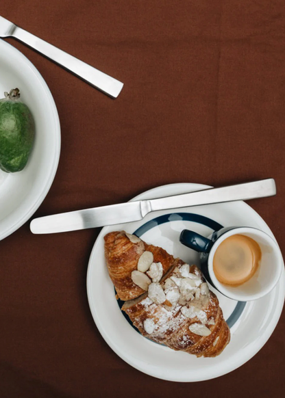 Café au lait with a croissant on a white plate on a brown tablecloth.