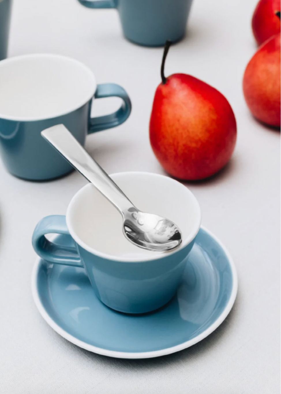 Blue cup with saucer and spoon on a white surface with apples in the background