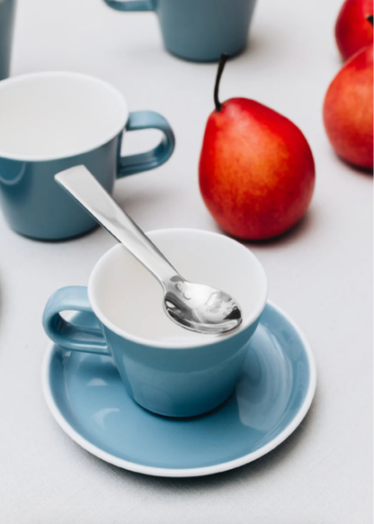 Blue cup with saucer and spoon on a white surface with apples in the background