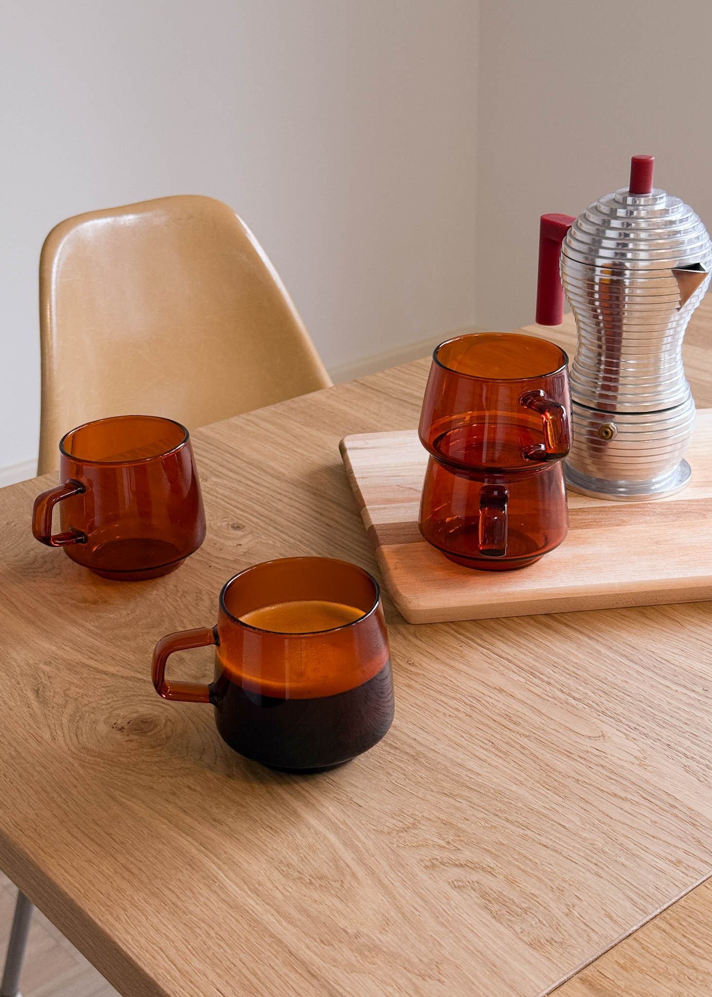 Three amber-colored glass mugs on a wooden table with a chair and a metallic pitcher in the background.