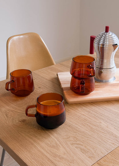 Three amber-colored glass mugs on a wooden table with a chair and a metallic pitcher in the background.