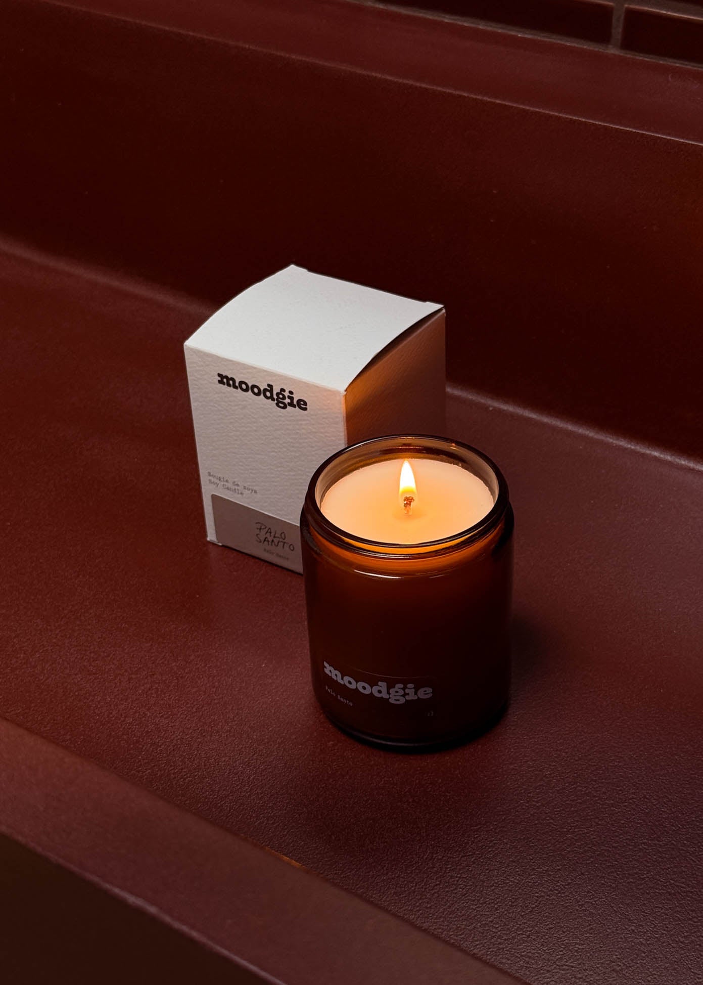 Moodgie candle with a lit flame on a dark surface next to its packaging box.