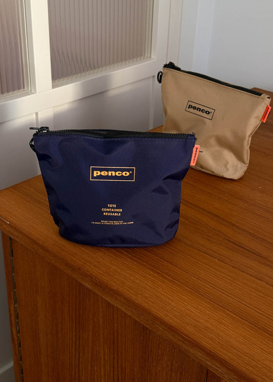 Navy and beige zipper pouches with 'penco' branding on a wooden surface.