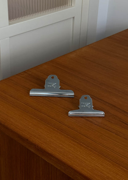 Two metal clips on a wooden surface with a white cabinet in the background.