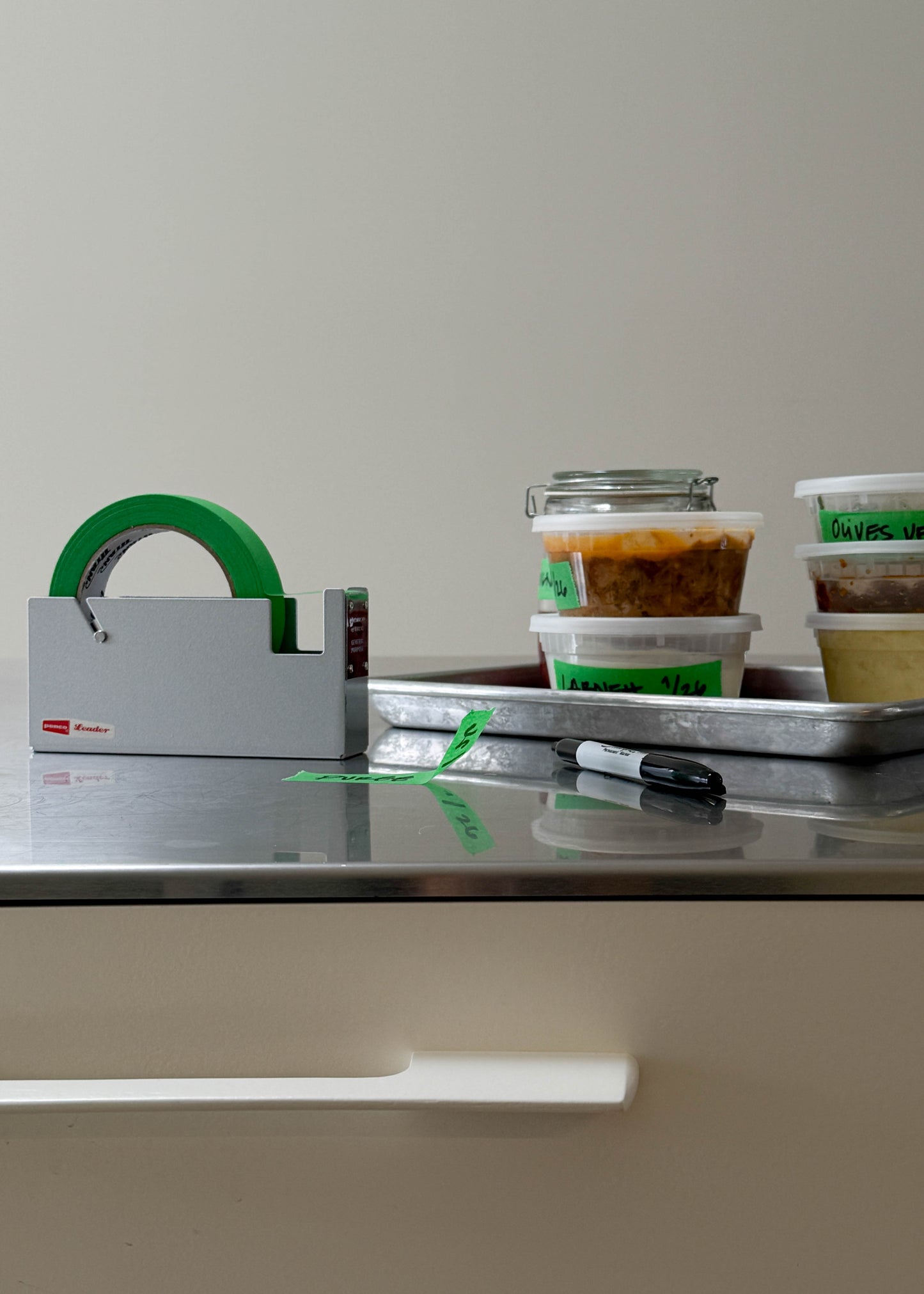 Stainless steel counter top with silver tape dispenser and green tape. Containers of food with green tape labels on them.