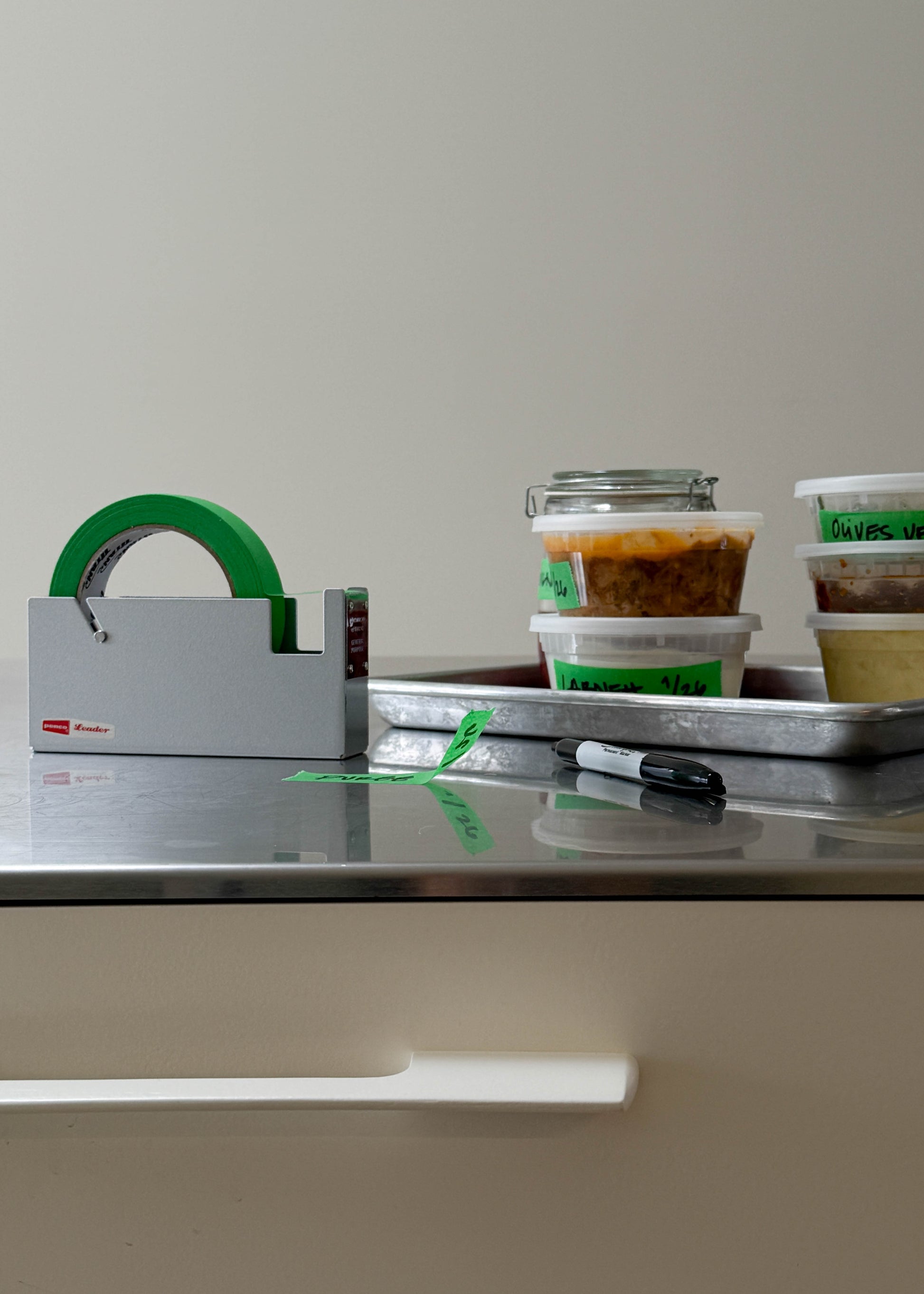 Stainless steel counter top with silver tape dispenser and green tape. Containers of food with green tape labels on them.