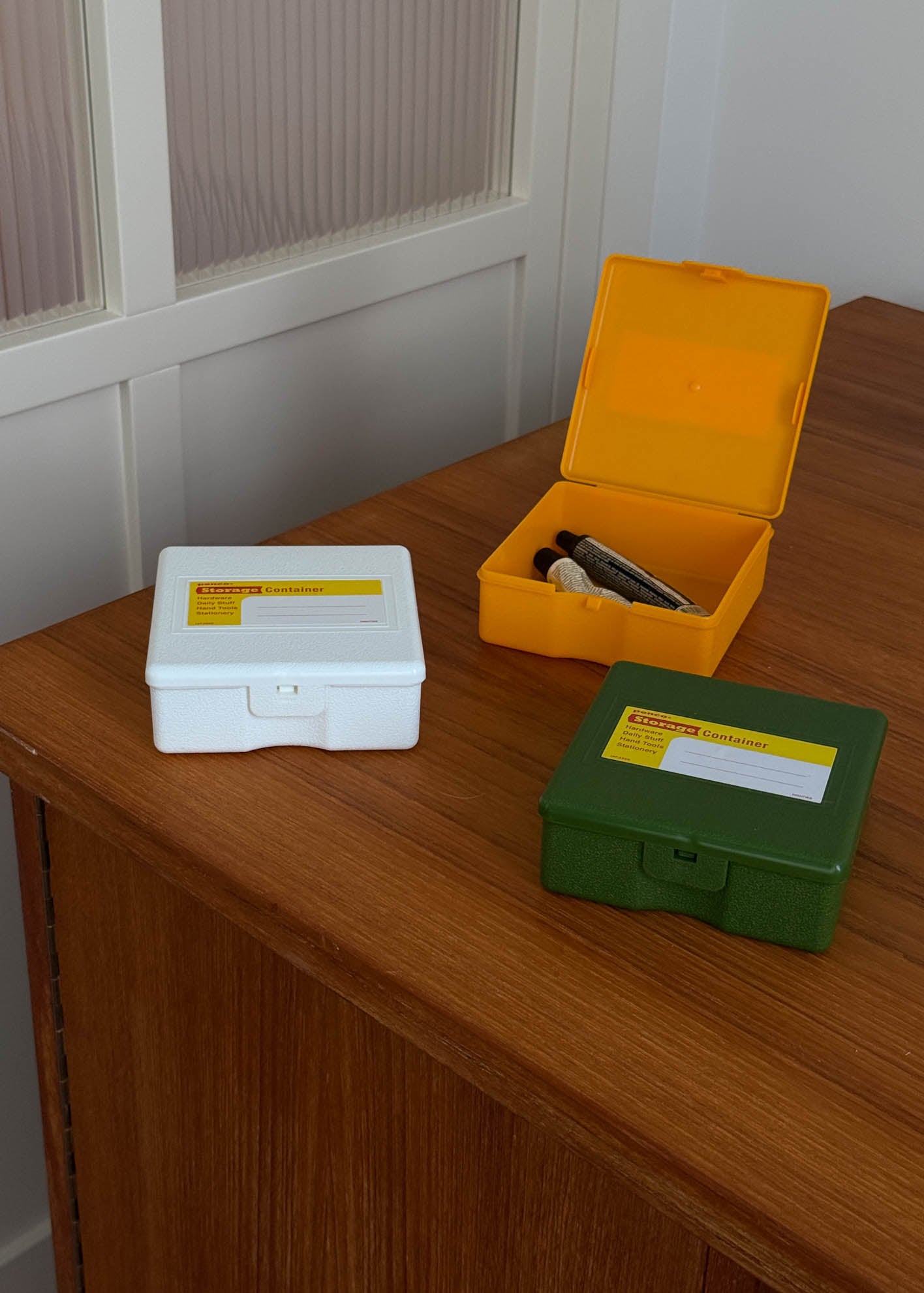 Three small plastic storage containers in white, yellow and green on a wooden surface with a white wall in the background