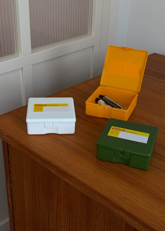 Three small plastic storage containers in white, yellow and green on a wooden surface with a white wall in the background