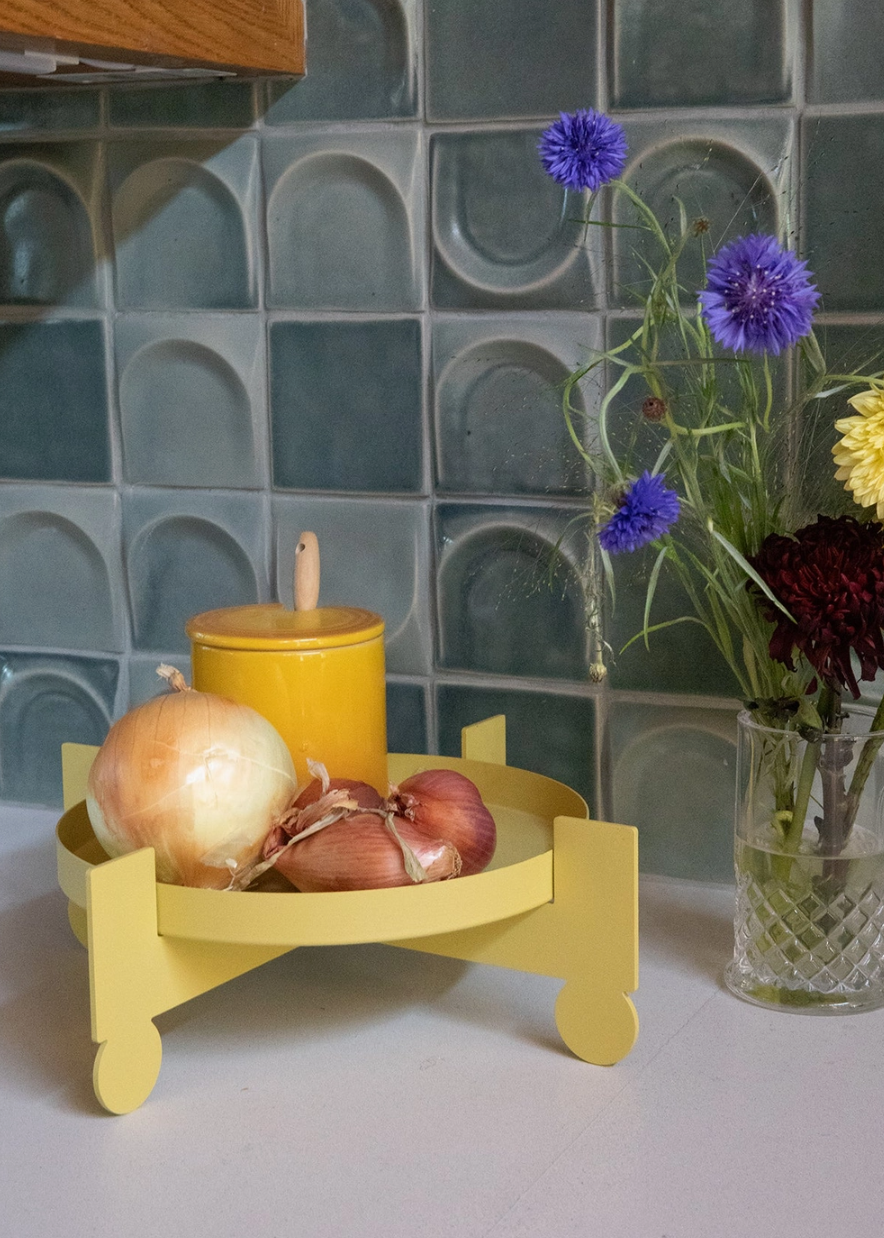 Yellow metal plant stand being used as a  tray with onions and a yellow container on a white kitchen counter infront of a tiled wall background