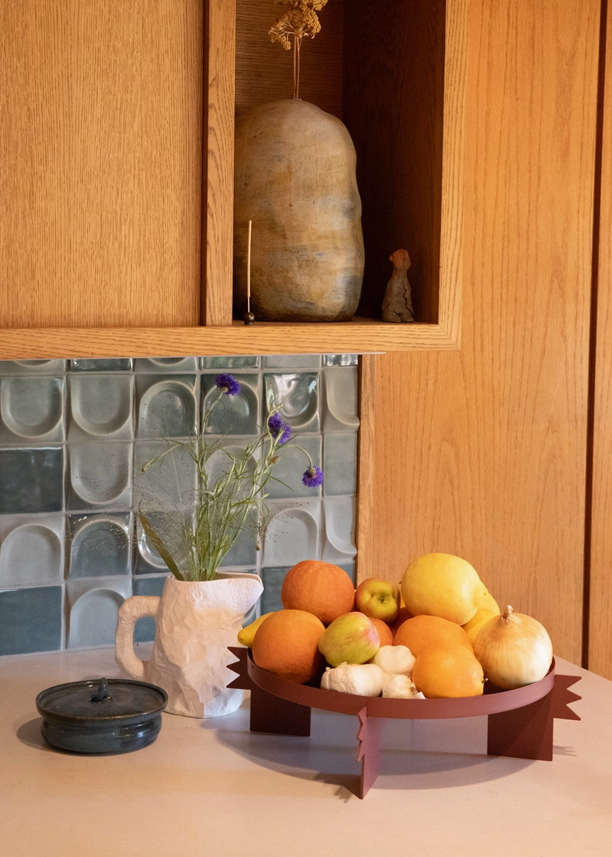 Geometric fruit stand with oranges, apples, and pears on a kitchen counter with wooden cabinets and a tiled backsplash.