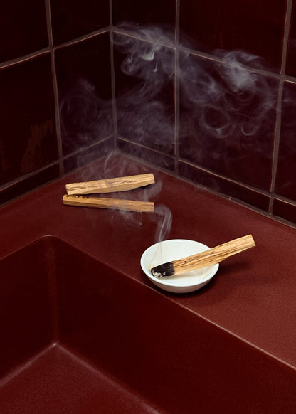 Palo santo sticks with smoke on a red surface against a tiled wall.